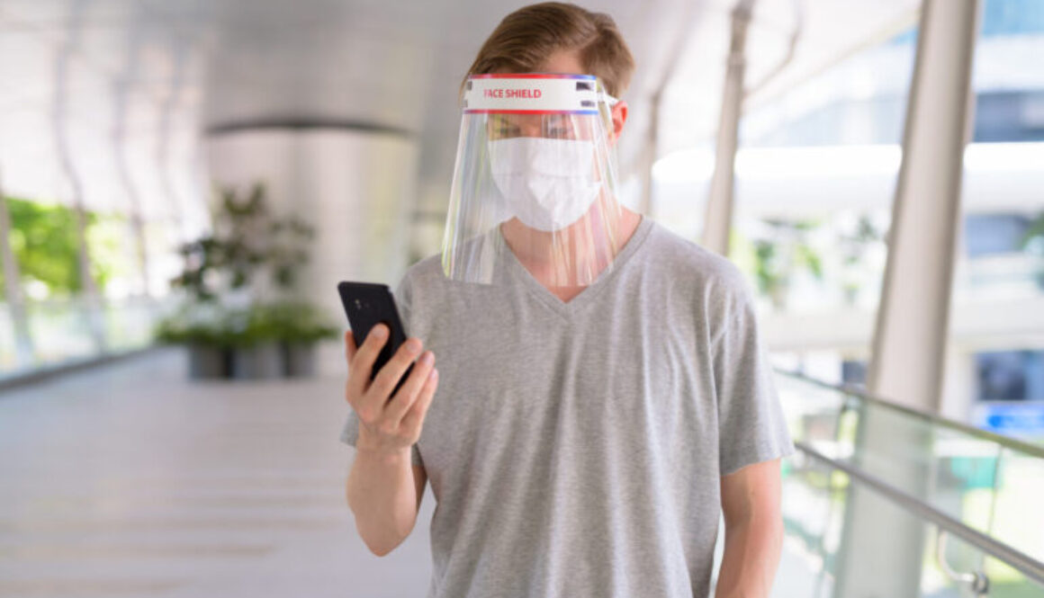 Young man using phone with mask and face shield for protection from corona virus outbreak in the city outdoors